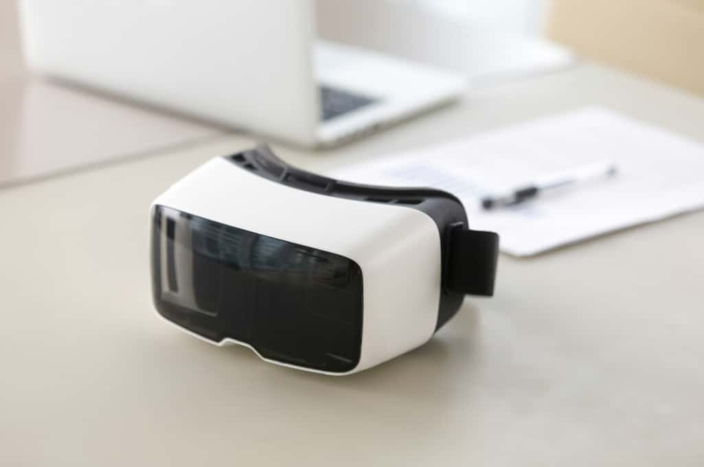 A VR headset rests on a desk beside a laptop and papers